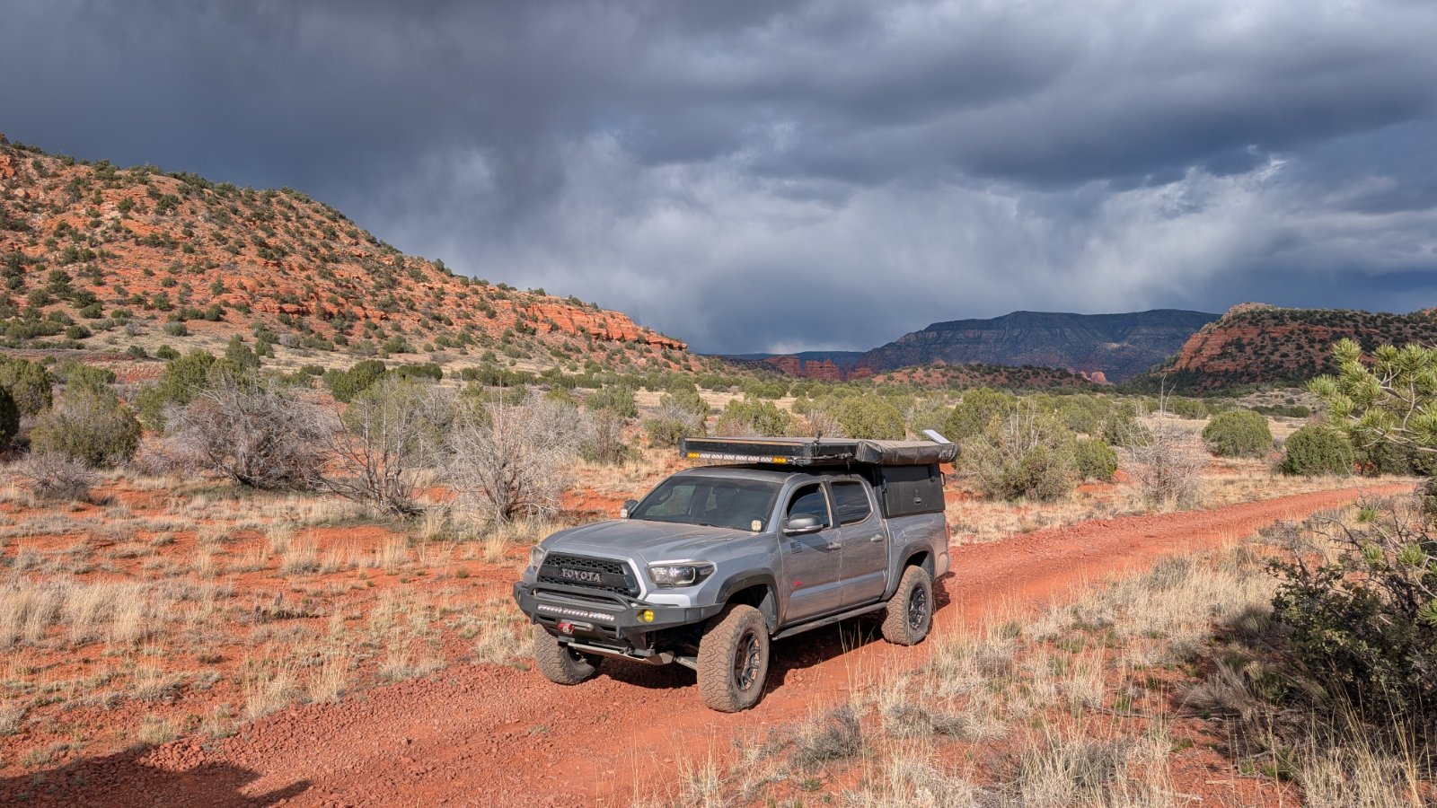 Silver Toyota Tacoma with roof rack and gear parked on red dirt in desert scrubland with red rock hills and storm clouds overhead