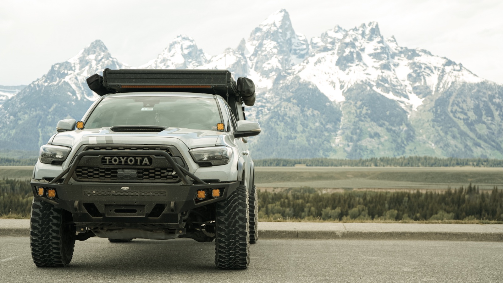 White Toyota Tacoma with roof rack and overland gear parked on asphalt with snow-capped mountain range and coniferous forest in background