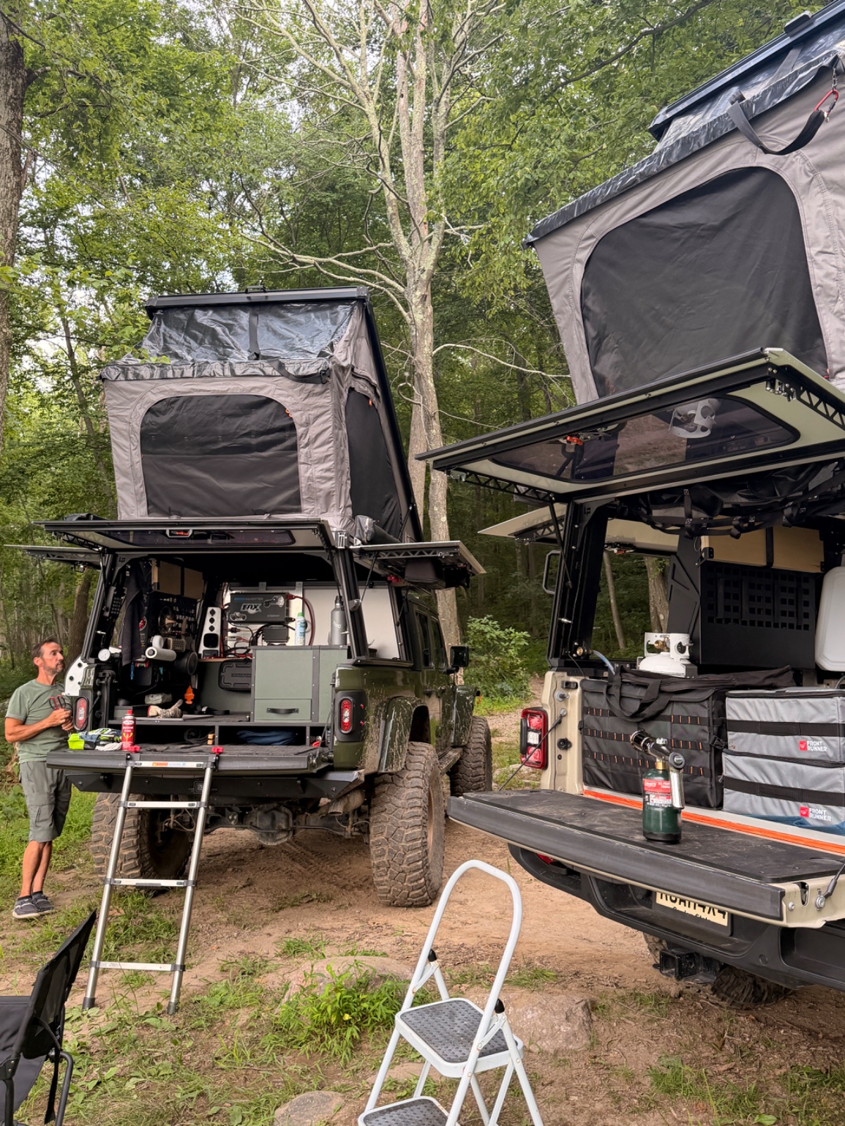 Two roof-top tent equipped off-road vehicles parked in a wooded campsite with camping gear and accessories visible, including a white step ladder and storage containers.