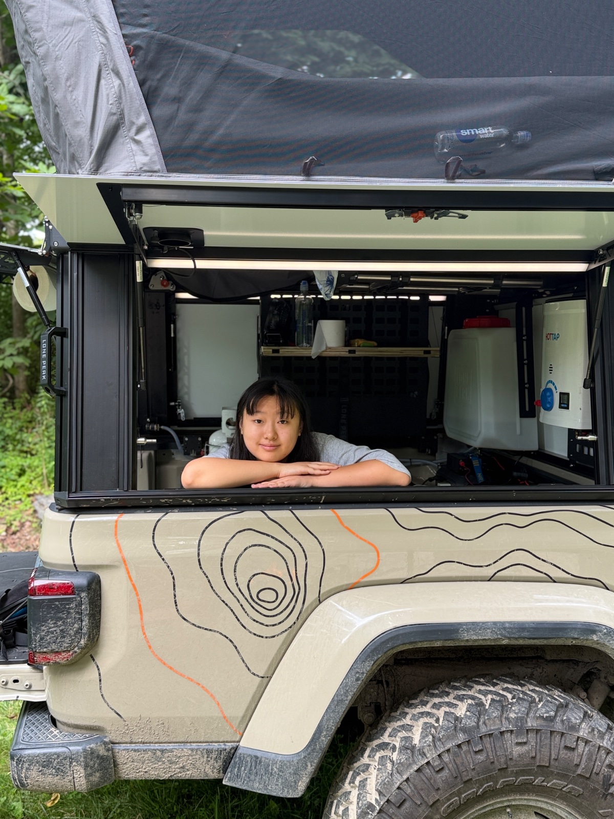 Young woman smiling from open window of cream-colored overlanding camper van with topographic map artwork, equipped with water heater and interior storage