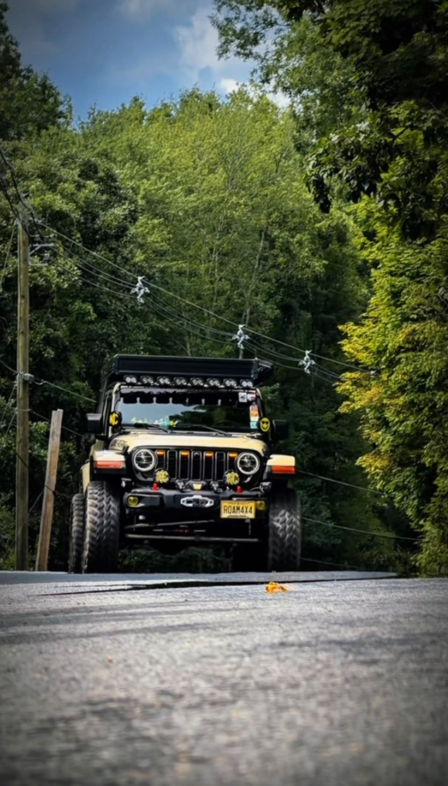 Modified black Jeep Wrangler with yellow accents driving under a low clearance bridge on a tree-lined rural road