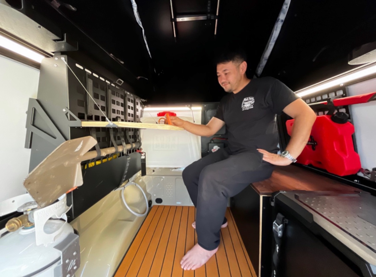 Man measuring interior of a boat cabin with a tape measure, showing the galley and storage compartments with wooden flooring and modern lighting.