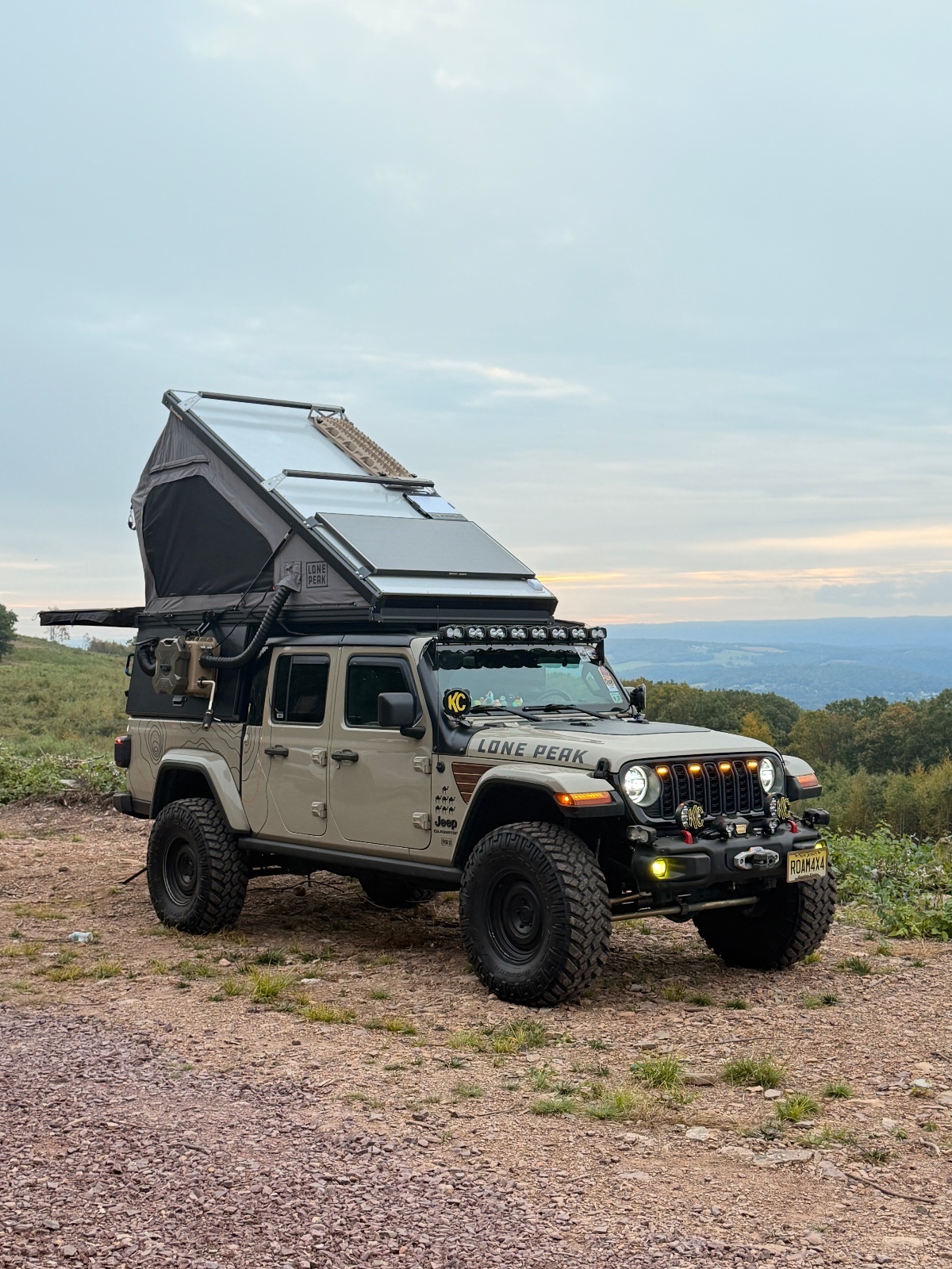 Tan Jeep Gladiator with roof rack and black rooftop tent parked on mountain overlook with valley view at dusk