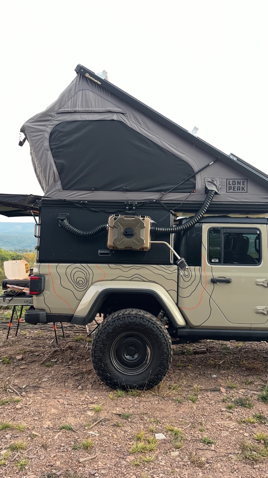 Tan Land Rover with roof-top tent deployed at a mountainous overlanding campsite, featuring topographic map decals and tan water container mounted on rear panel