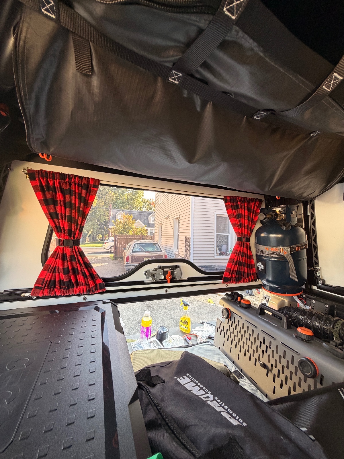 Interior view of a truck bed camper setup with red plaid curtains, black rubber flooring, storage containers, and gas equipment visible through the open canvas roof to a residential driveway
