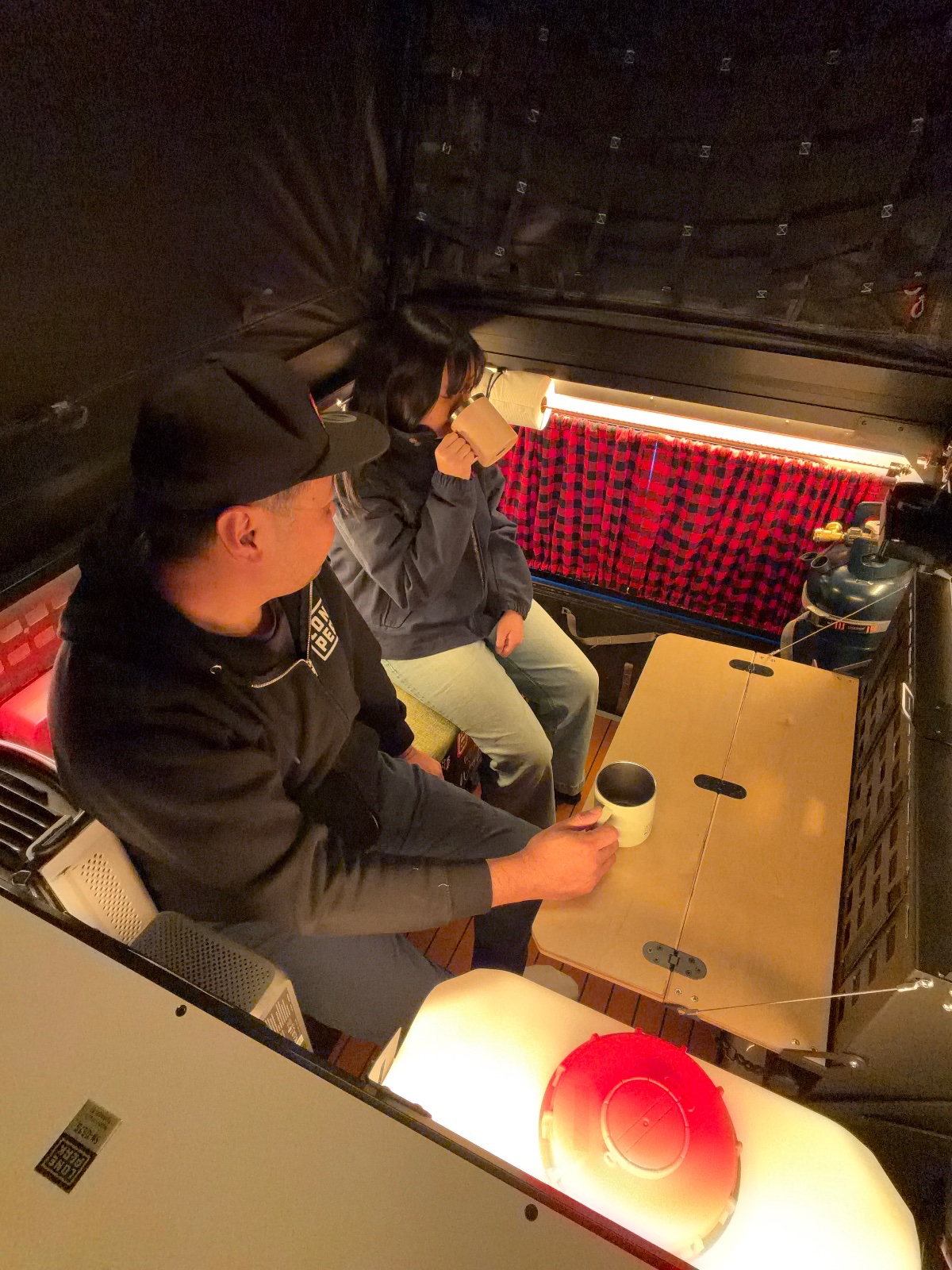 Two people sitting in the back of a vehicle at night, examining a red plaid blanket and cardboard box with camping gear and red LED lights visible