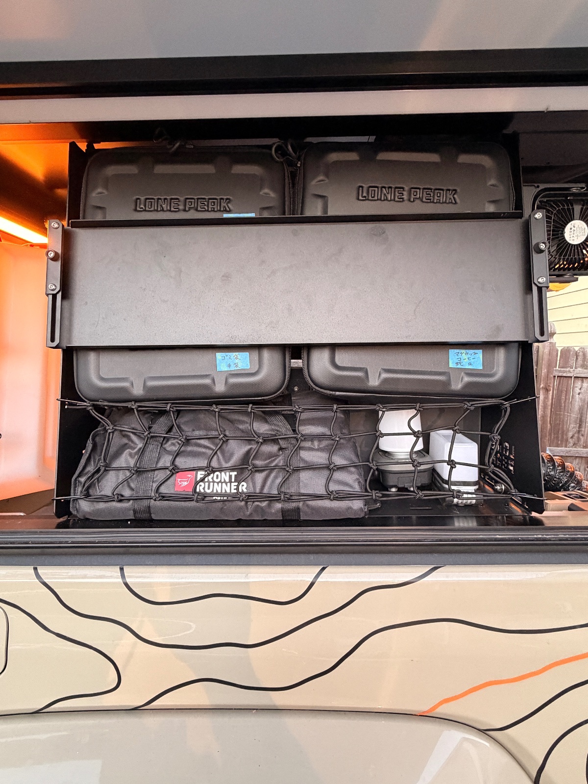 Roof-mounted storage setup on overlanding vehicle featuring Lone Peak hard cases, Front Runner gear basket with bungee net, and cooling fan for expedition camping