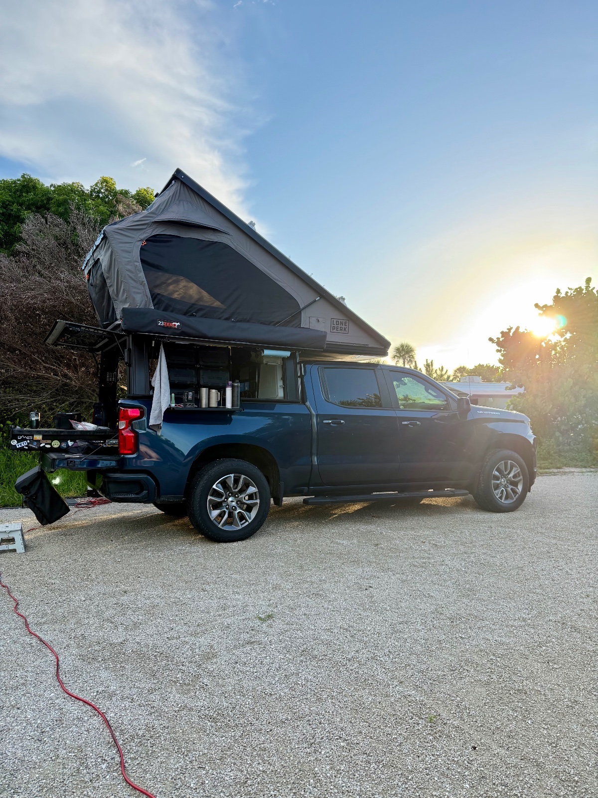 Blue pickup truck with roof-top tent deployed at a paved campground during golden hour sunset