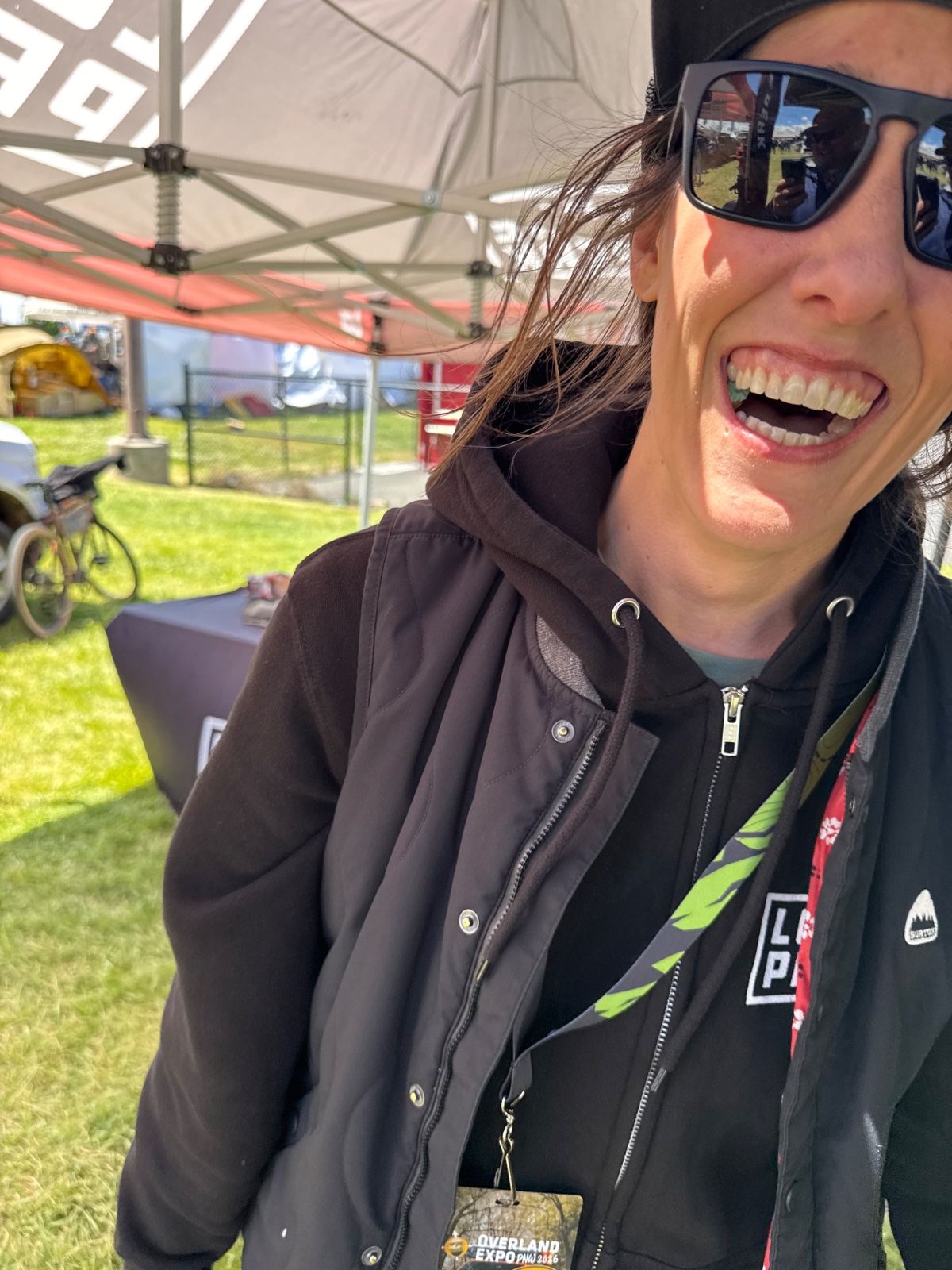 Woman wearing sunglasses and black jacket with lanyard at outdoor event under tent canopy with green grass and vehicles visible in background