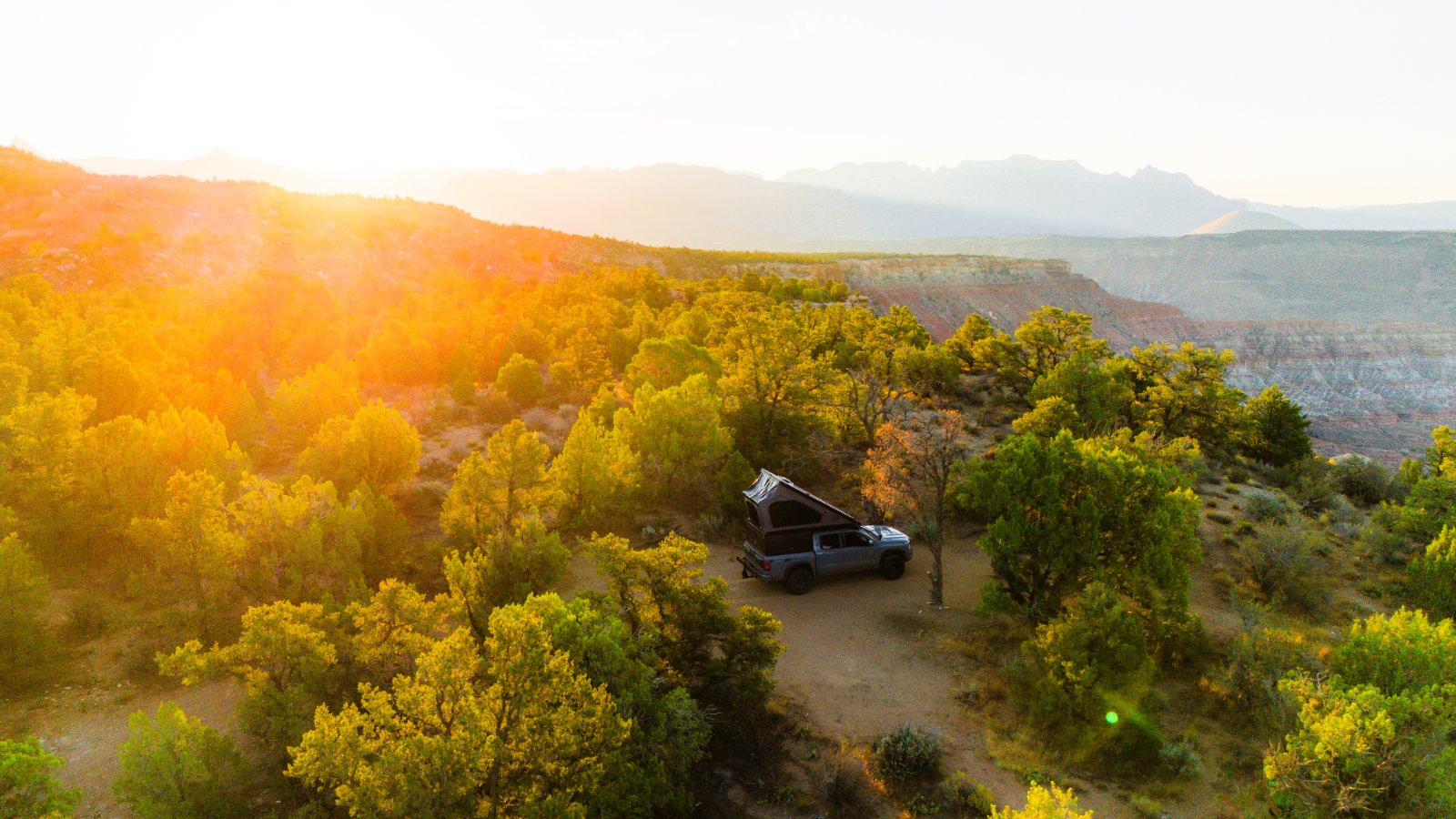 A pickup truck with an open camper shell parked among juniper and pinyon pine trees at sunrise, overlooking a desert canyon landscape with distant plateaus