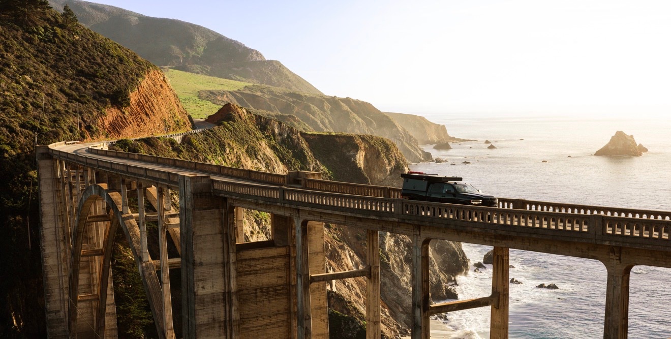Black SUV parked on a dramatic coastal bridge overlooking rugged cliffs, green hillsides, and rocky outcrops along a misty Pacific coastline at golden hour