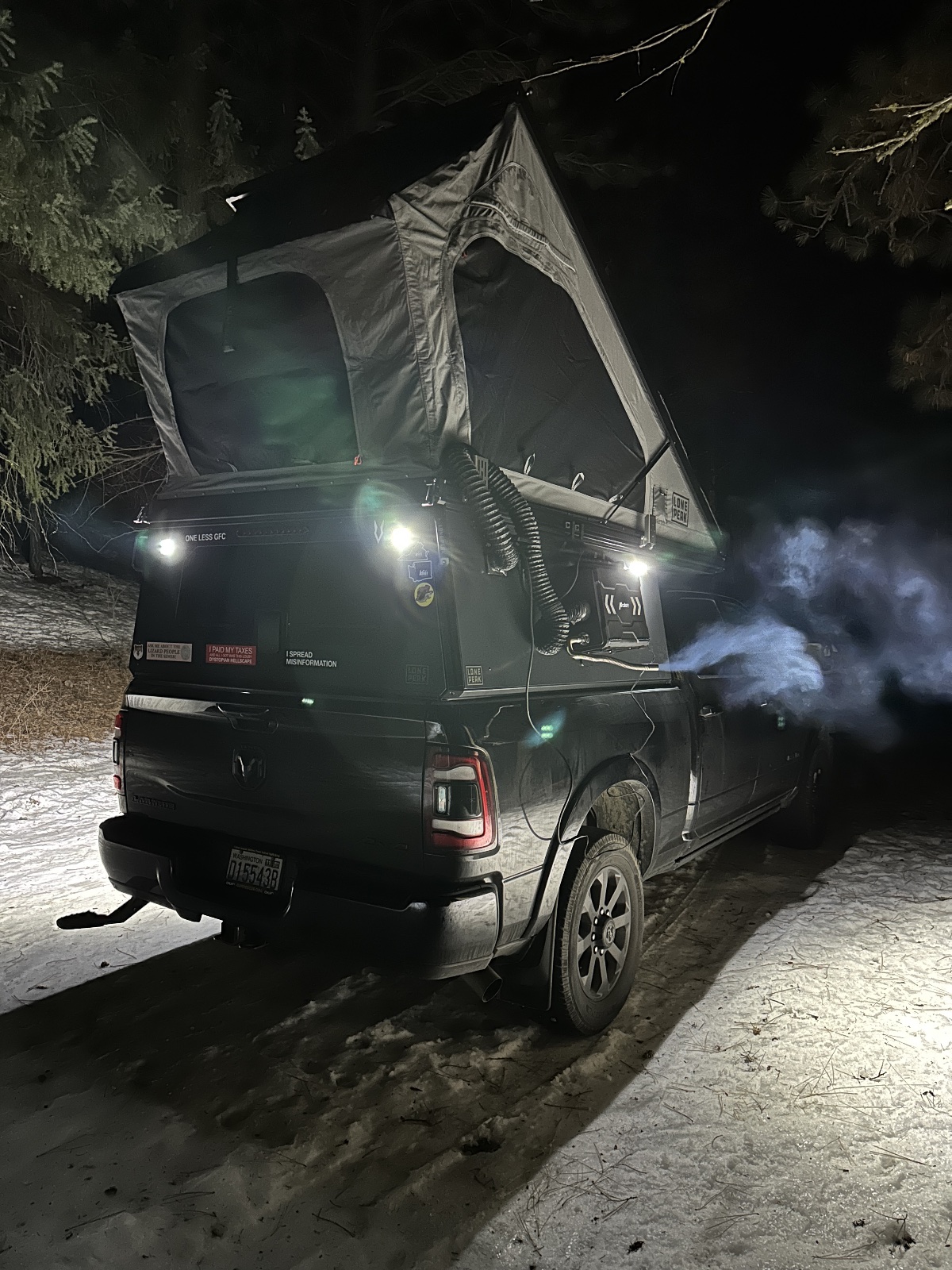 Black pickup truck with rooftop tent set up at night in a forested campsite, illuminated by LED lights and a misting system creating atmospheric vapor