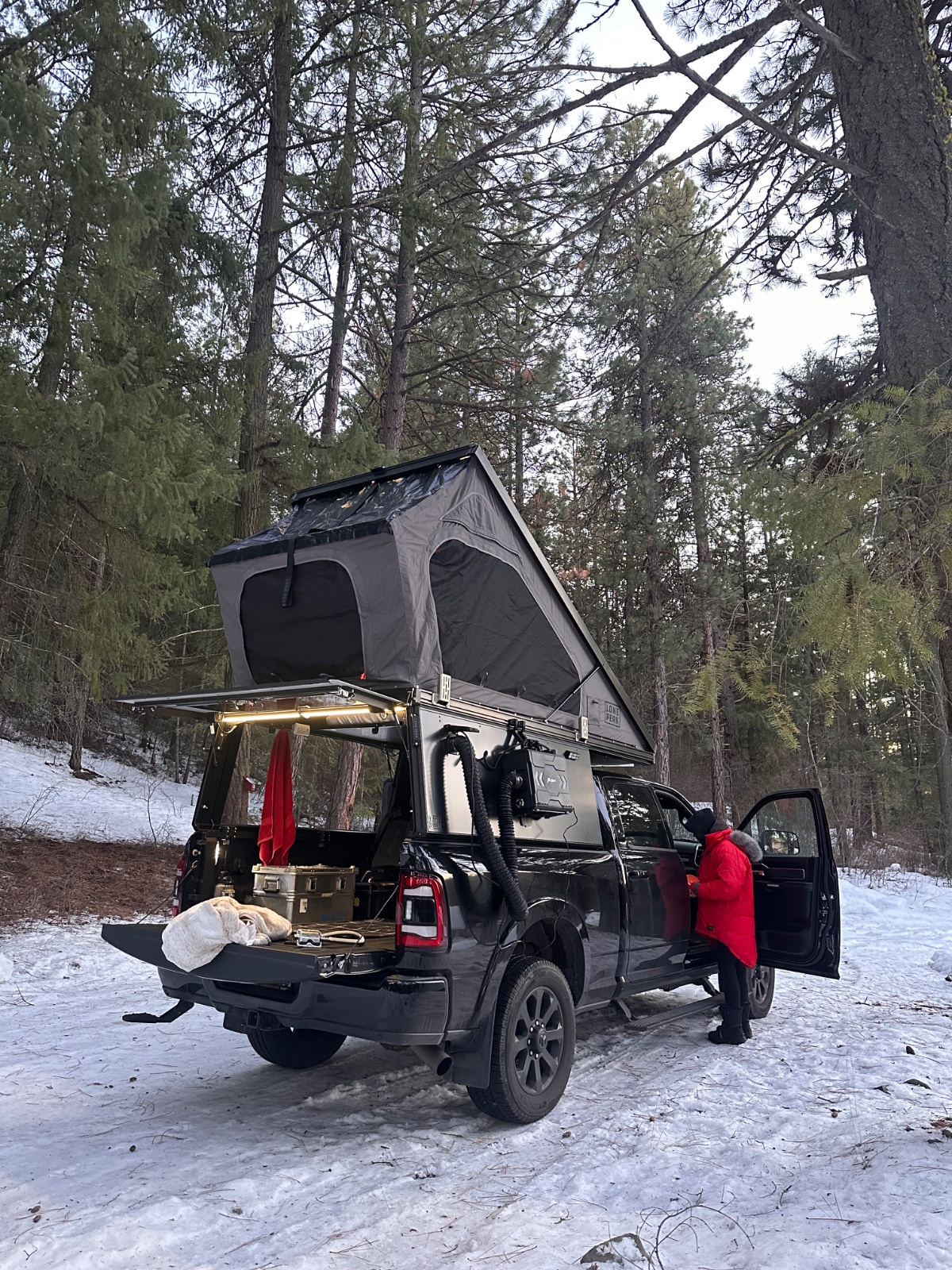 Black vehicle with roof tent deployed in a snowy forest campsite, person in red jacket standing beside open cargo area with camping gear