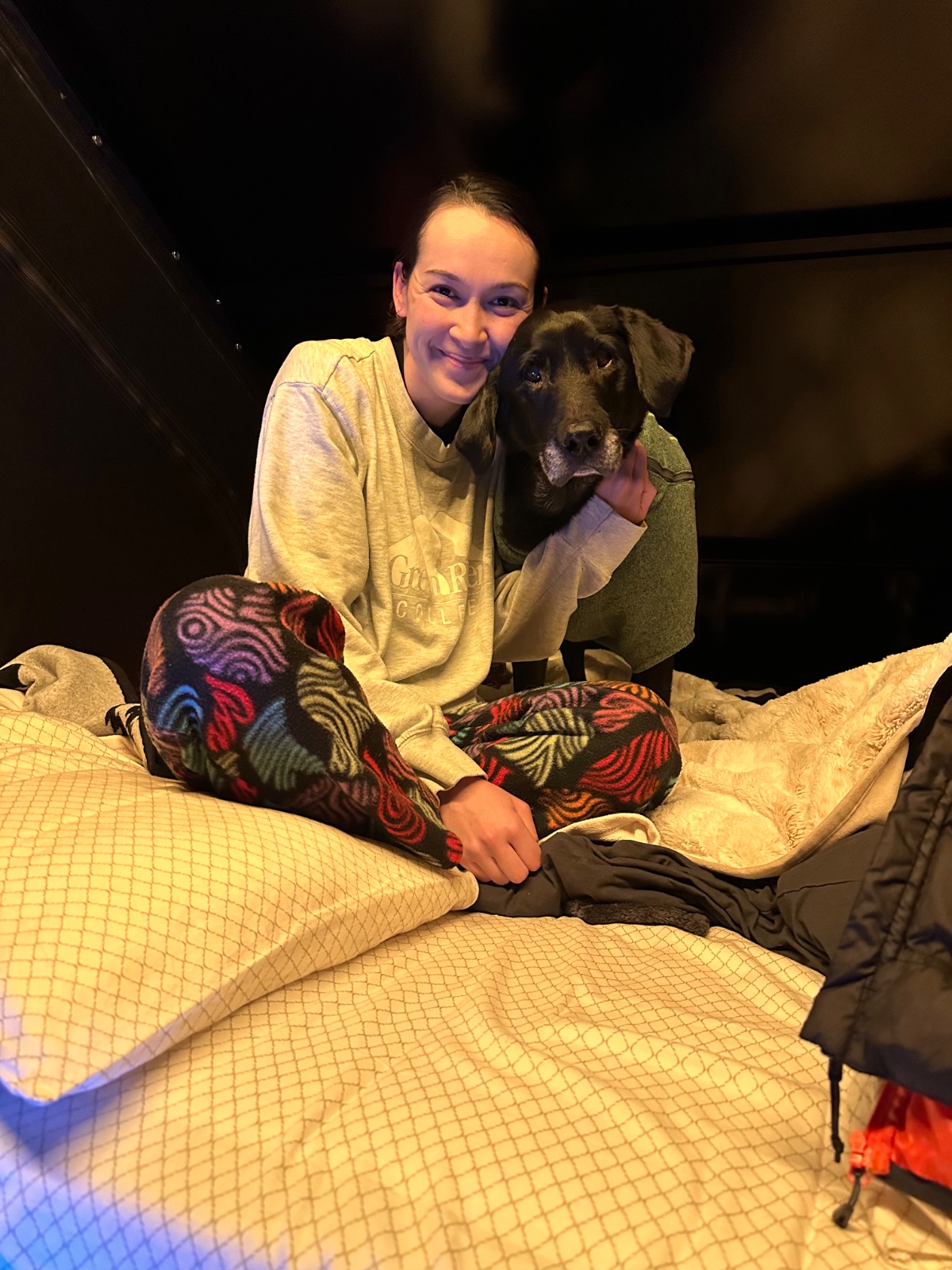Young woman smiling while sitting on a bed with a black dog with gray muzzle, wearing colorful patterned leggings and casual shirt