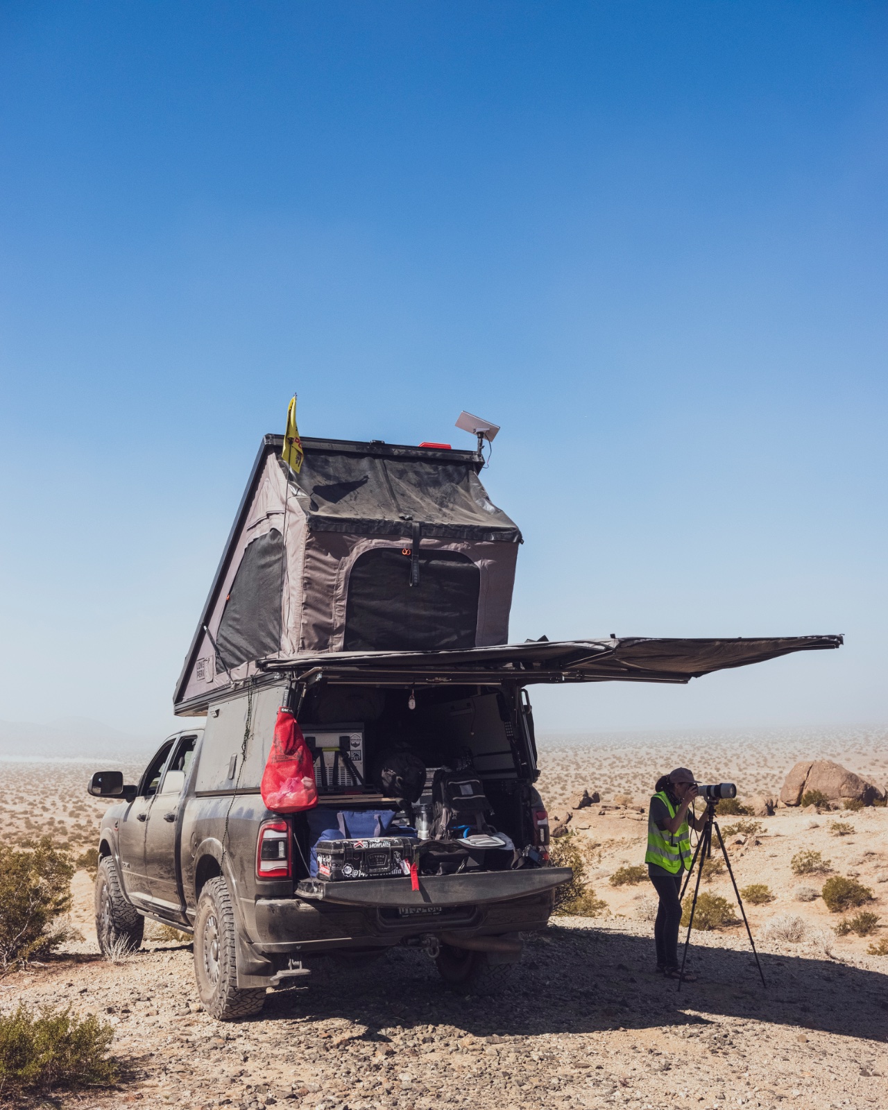 A truck with a roof tent and awning set up in a desert landscape with a person operating a surveying tripod nearby