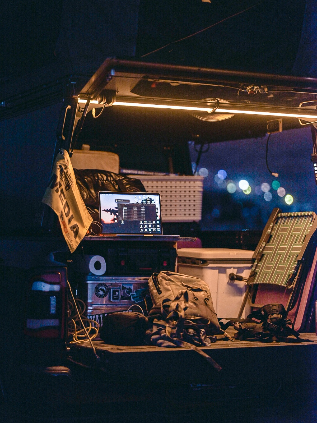 Van interior at night with laptop displaying map application, camping gear, folding chair, and ambient LED lighting overhead