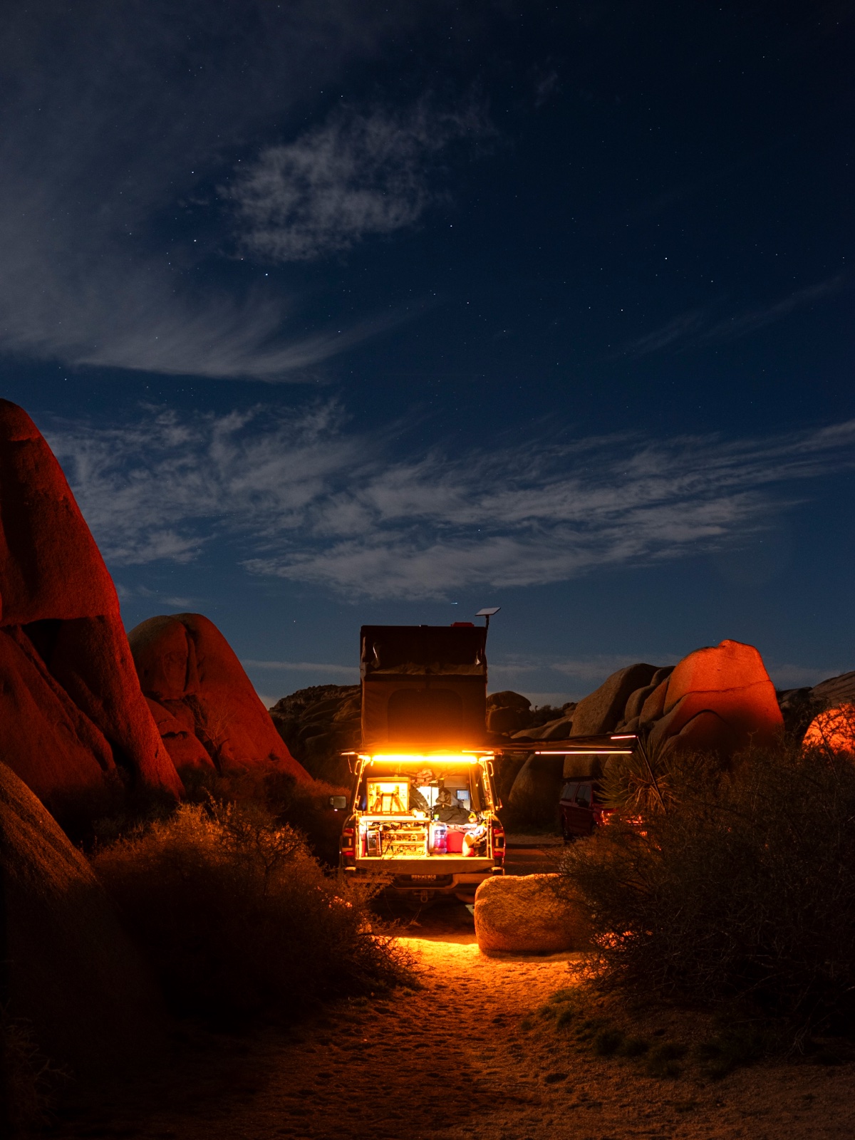 Illuminated pickup truck camper setup nestled among large boulders in desert terrain under starry night sky