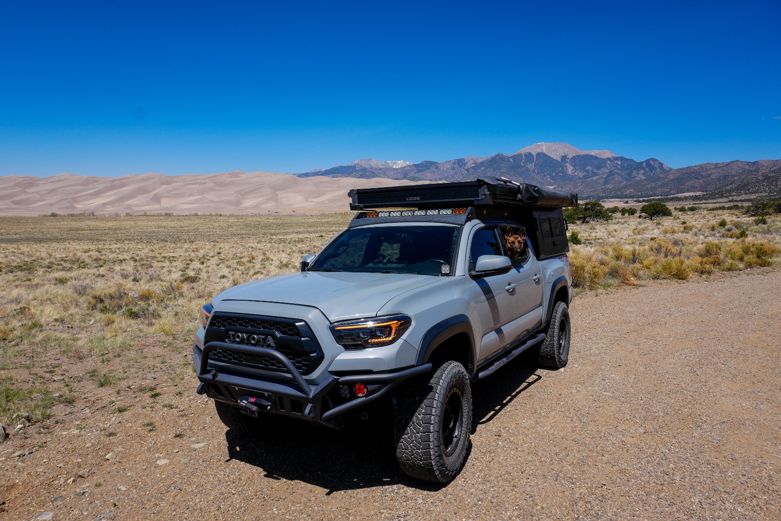 White Toyota Tacoma with roof rack and front bumper guard parked on desert terrain with sand dunes and mountains in background under clear blue sky