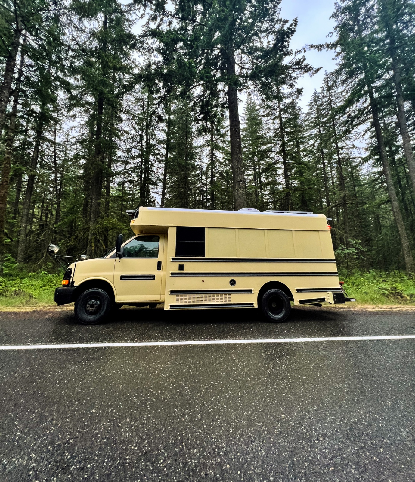 Yellow custom truck camper parked on asphalt road with tall coniferous forest backdrop