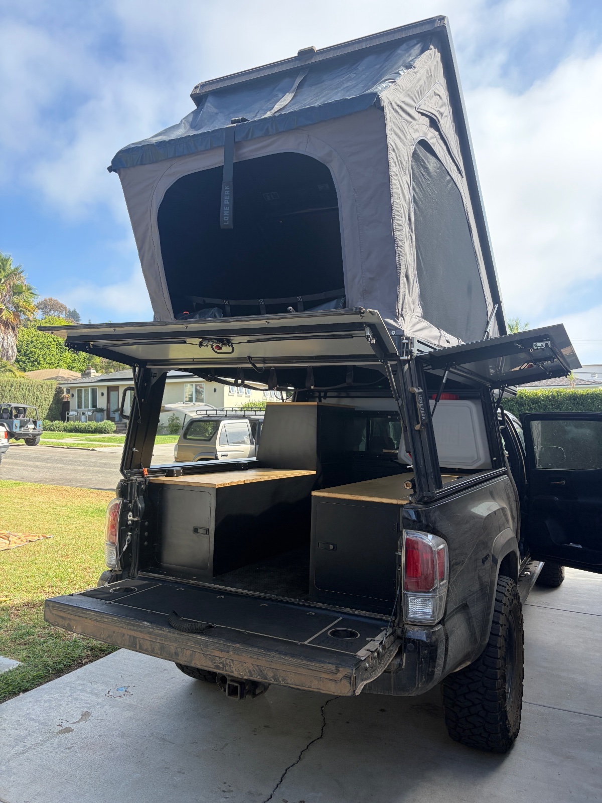 Black pickup truck with roof tent fully extended and wooden storage boxes, parked on residential driveway with palm trees visible in background