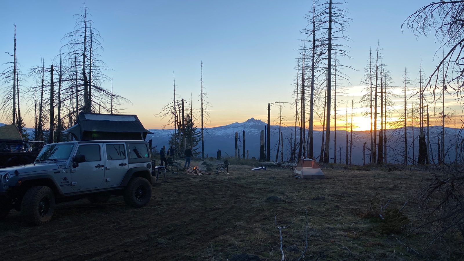 White Jeep Wrangler with roof tent parked at alpine campsite surrounded by dead trees at sunset with snow-capped mountain peak in distance