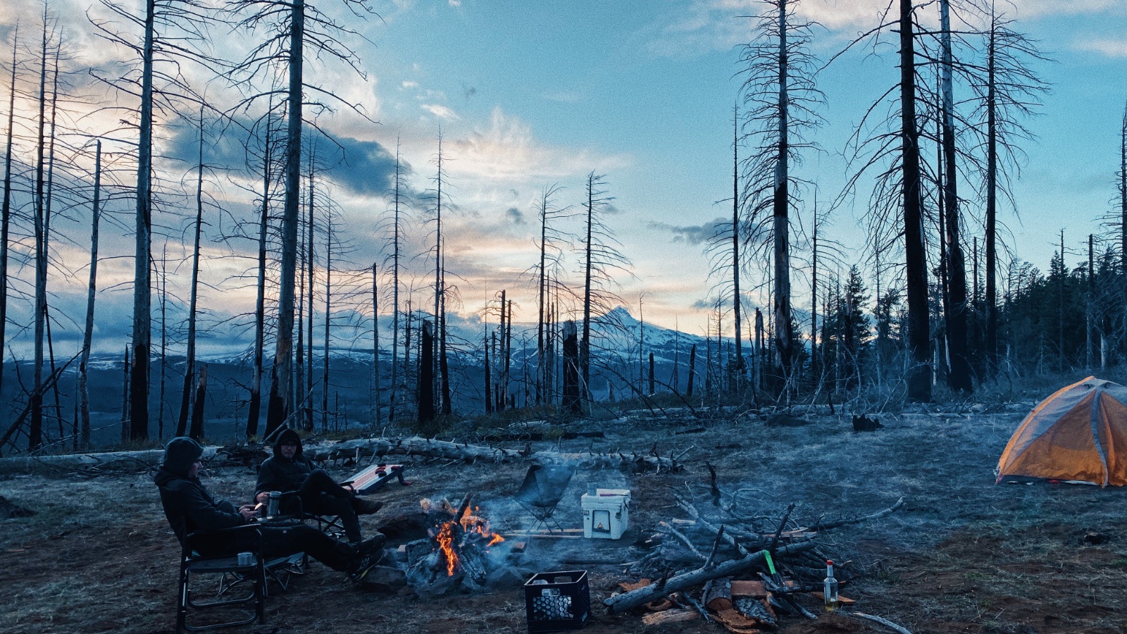 Two campers sit by a campfire in a burned forest landscape with mountains visible at dusk, with an orange tent and camping gear nearby