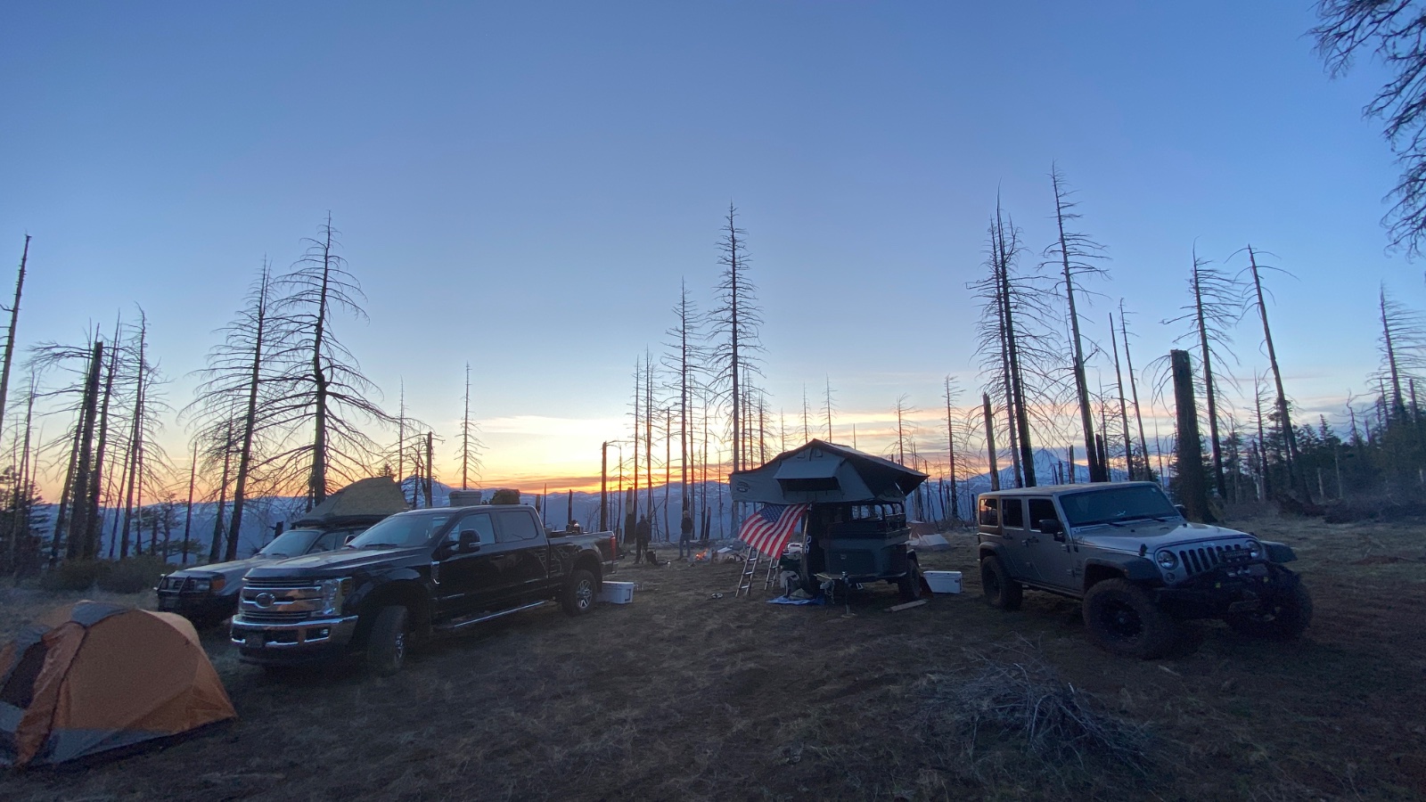 Overlanding camp at sunrise with Chevrolet trucks, Jeep Wrangler, tent, roof-top tent shelter, and American flag surrounded by dead lodgepole pines in high alpine terrain