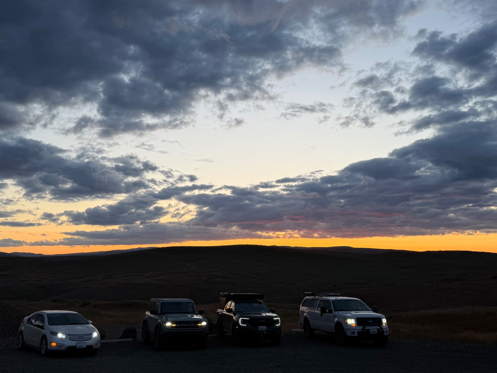 Four vehicles including a white sedan, dark SUVs, and a white truck lined up on a desert overlook at sunset with storm clouds and golden sky