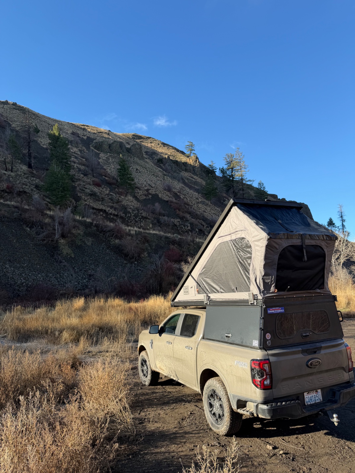 Cream-colored Ford F-150 pickup truck with pop-up roof tent deployed, parked on desert scrubland below rocky hillside with scattered evergreen trees and clear blue sky